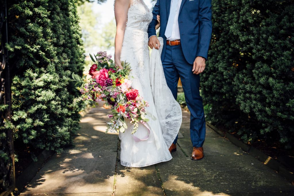 Bride & Groom holding pink bridal bouquet, lace wedding dress, blue suit with brown belt and shoes